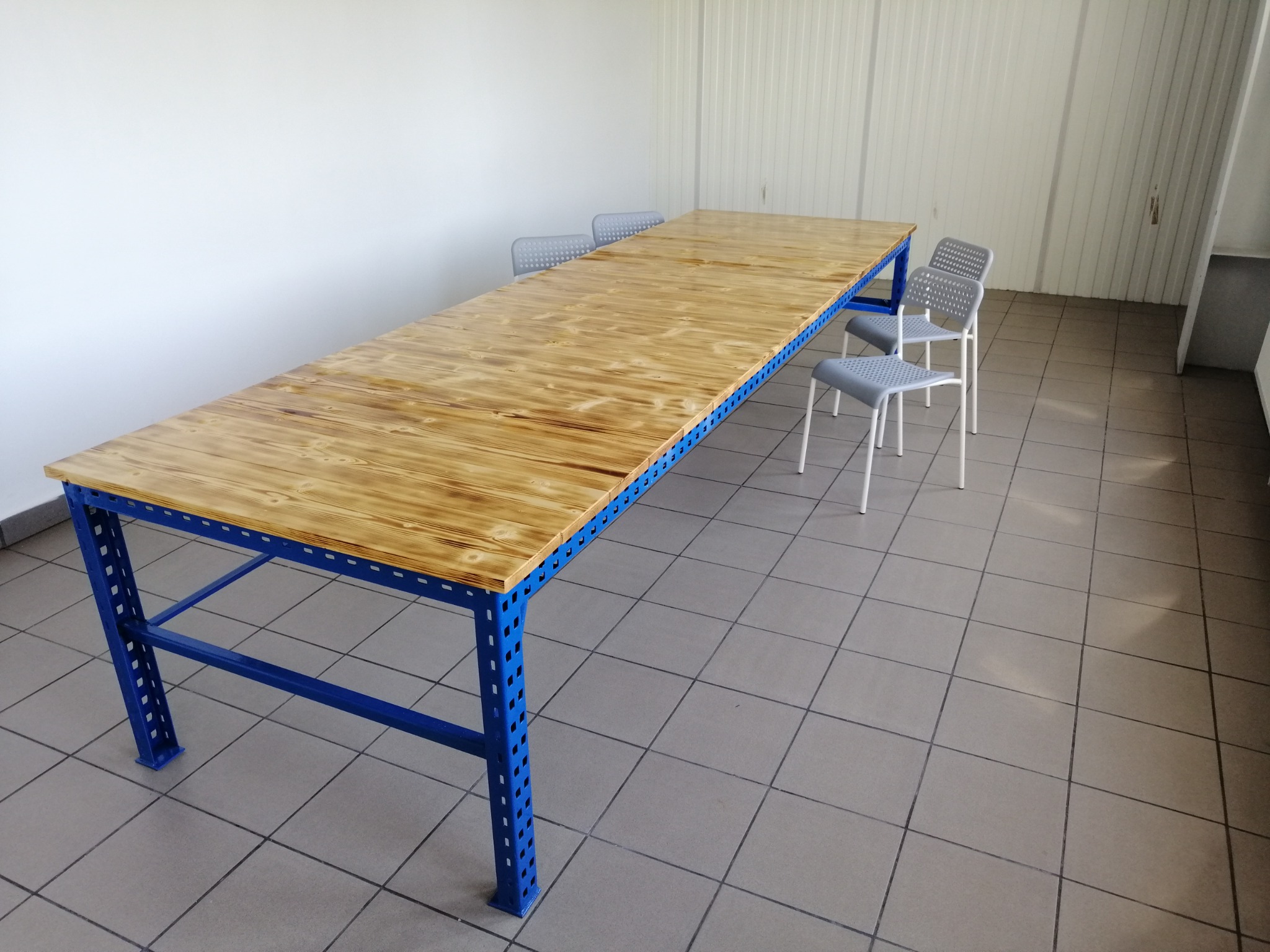 Długi stół warsztatowy z blatem z jasnego drewna i niebieską, perforowaną metalową konstrukcją, stoi na szarej podłodze z płytek; w tle białe ściany i krzesła.
