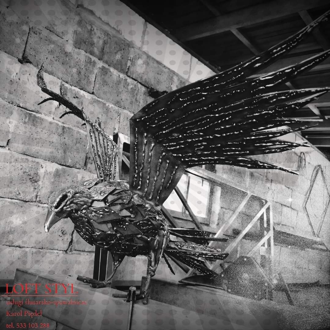 Czarno-białe zdjęcie metalowej rzeźby orła z rozpostartymi skrzydłami, wykonanej z pospawanych elementów, na tle surowej ściany z cegieł i metalowej konstrukcji schodów.