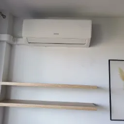 Biały klimatyzator Sinclair na ścianie, z instalacją w maskownicy. Dwie półki z jasnego drewna i obraz w czarnej ramie z motywem roślinnym dopełniają wystrój.