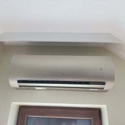 INSTALACJE Zabrze-Gliwice - Zamontowany klimatyzator w jasnym wnętrzu, widoczny pod sufitem, z dekoracyjną listwą. Ściana w delikatny wzór, dół kadru okno z brązową ramą.