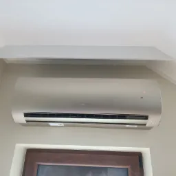 Zamontowany klimatyzator w jasnym wnętrzu, widoczny pod sufitem, z dekoracyjną listwą. Ściana w delikatny wzór, dół kadru okno z brązową ramą.