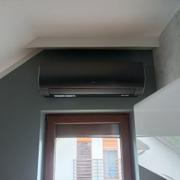 INSTALACJE Zabrze-Gliwice - Czarny klimatyzator ścienny zamontowany nad oknem z brązową ramą w szaro-białym wnętrzu domu. Widoczne elementy zabudowy.