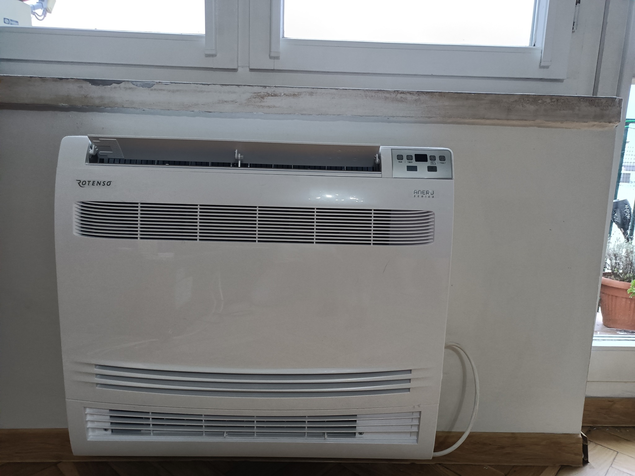 Biały klimatyzator Rotenso Aneri J Series, stojący przy ścianie z oknem, widoczne przewody. Montaż domowej klimatyzacji.