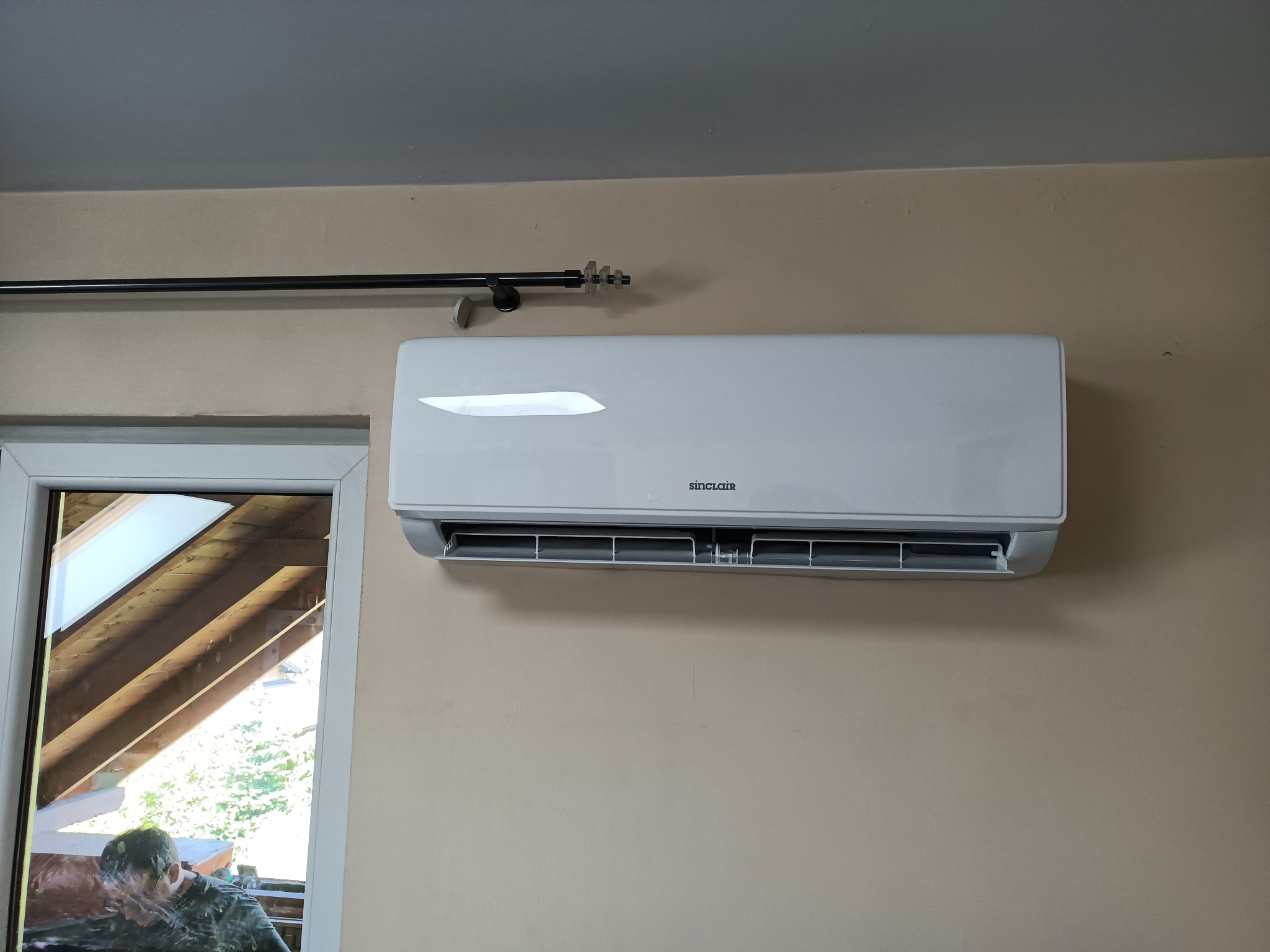 Biały klimatyzator Sinclair zamontowany na beżowej ścianie, widoczny fragment okna i karnisz. Domowa instalacja klimatyzacji.