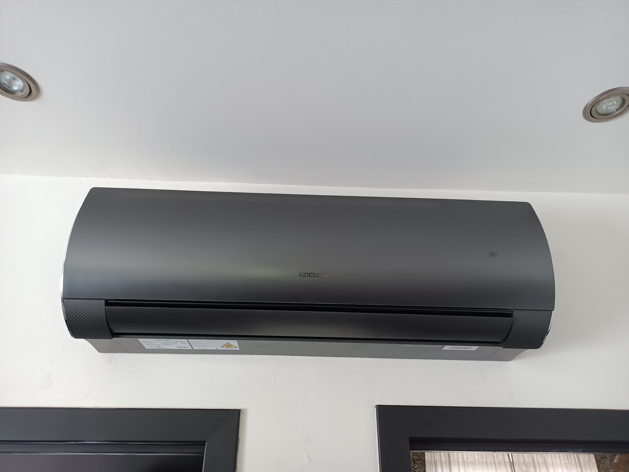 Czarny klimatyzator Sinclair zamontowany na białej ścianie, widoczne dwa reflektory sufitowe i fragmenty dwóch obrazów w czarnych ramach.