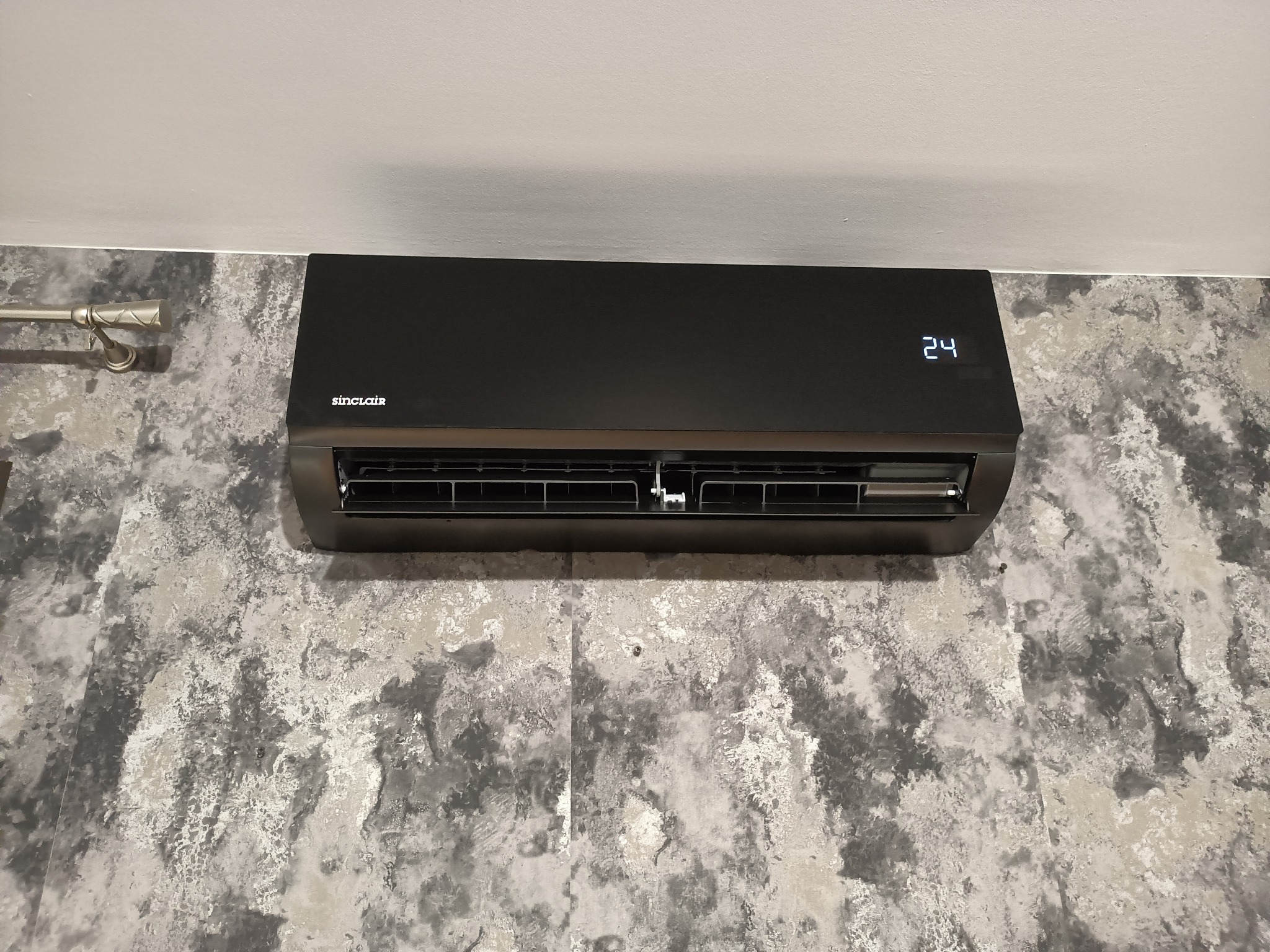 Czarny klimatyzator Sinclair z wyświetlaczem temperatury (24°C) zamontowany na ścianie o teksturze imitującej kamień. Widoczne elementy instalacji.