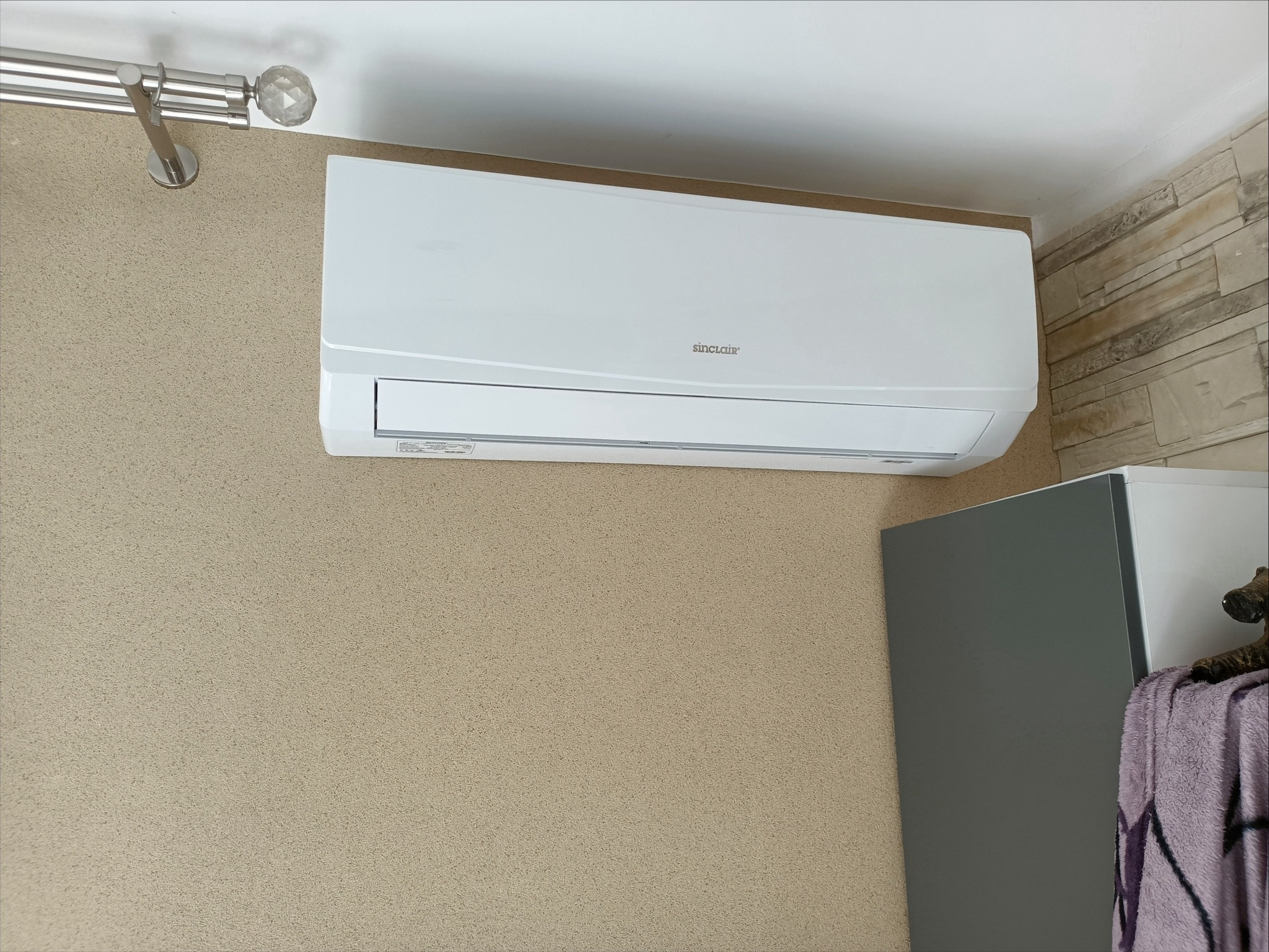 Biała klimatyzacja Sinclair zamontowana na ścianie o beżowej fakturze, z fragmentem karnisza i szafki w tle. Widok z dołu ukazuje detale urządzenia.