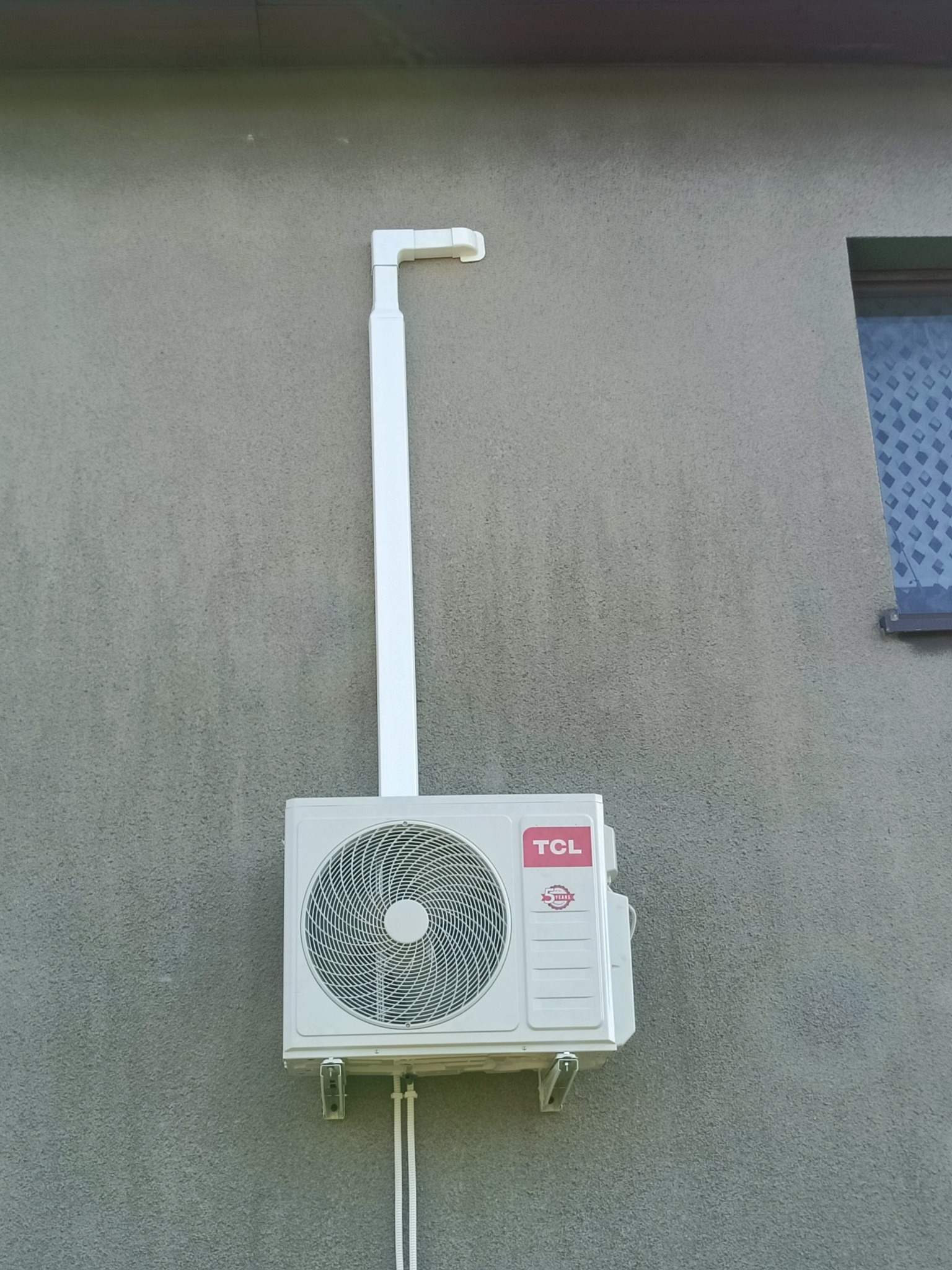 Zamontowana jednostka zewnętrzna klimatyzacji TCL na szarej elewacji budynku, z widoczną instalacją odprowadzającą, okno w tle. Widok z dołu.