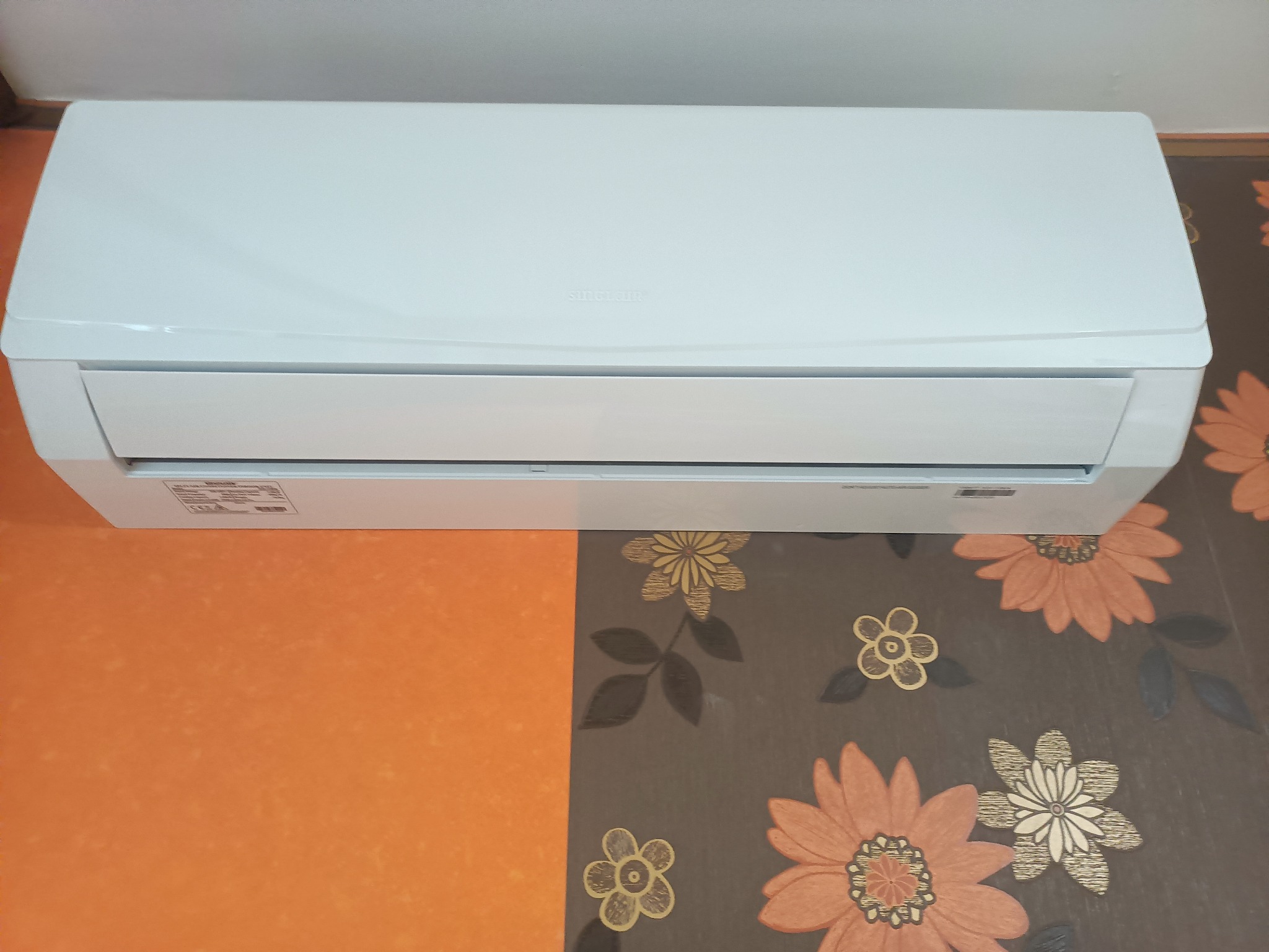 Biały klimatyzator Sinclair na tle wzorzystej tapety. Widoczny z przodu, zamontowany na ścianie.