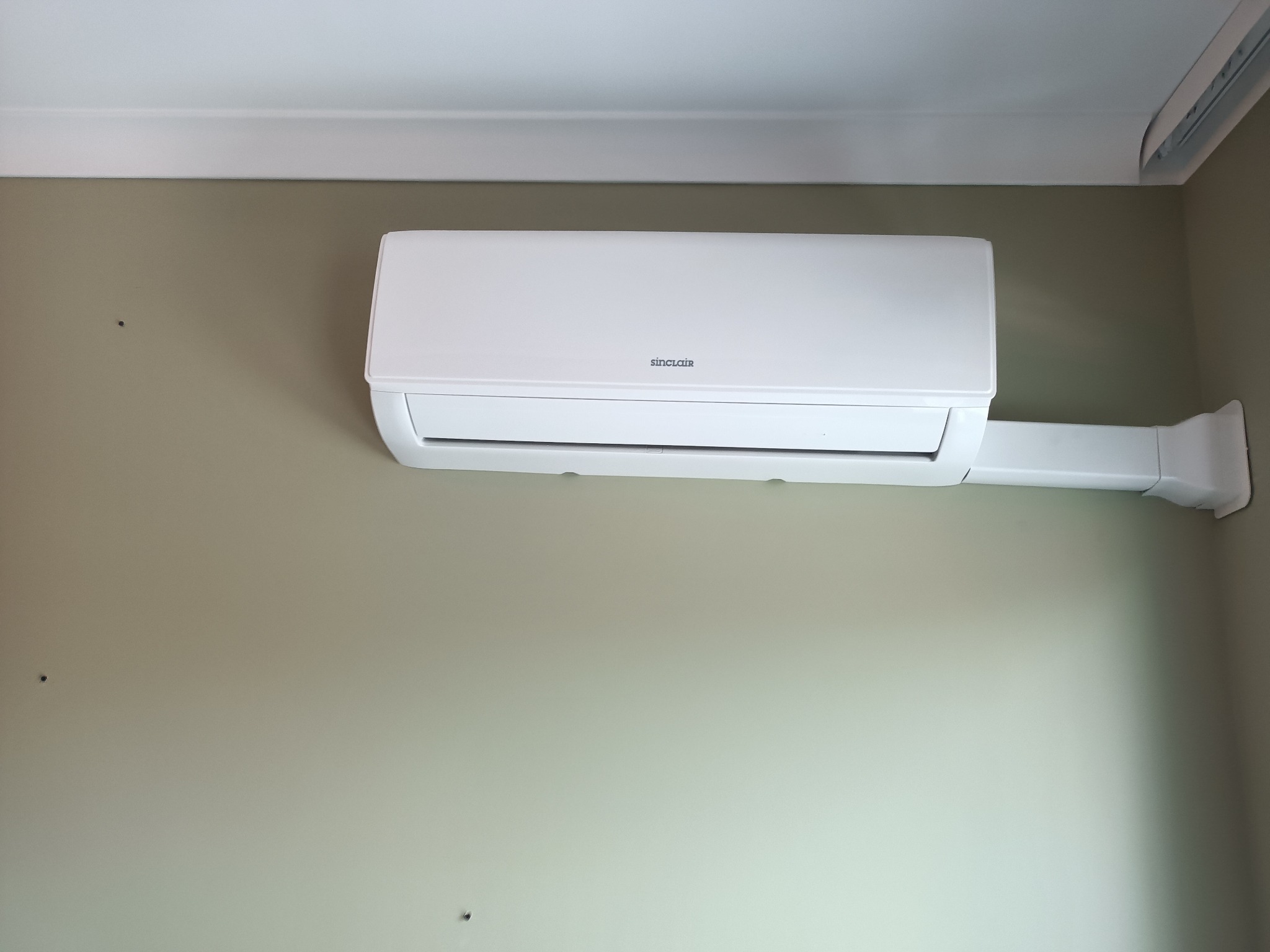 Biały klimatyzator Sinclair zamontowany na ścianie w jasnym pomieszczeniu, widoczny przewód odprowadzający, minimalistyczny design.