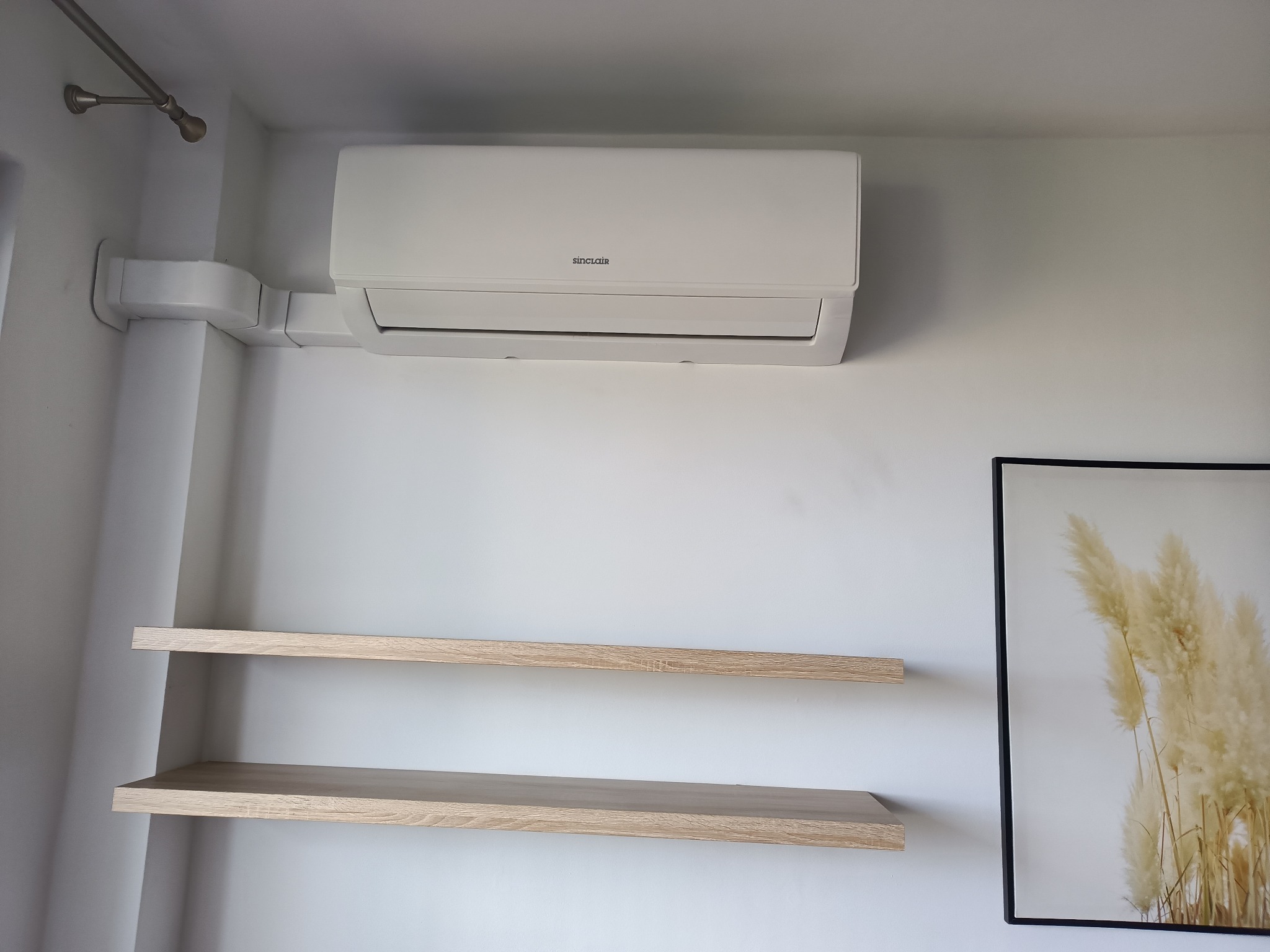 Biały klimatyzator Sinclair na ścianie, z instalacją w maskownicy. Dwie półki z jasnego drewna i obraz w czarnej ramie z motywem roślinnym dopełniają wystrój.