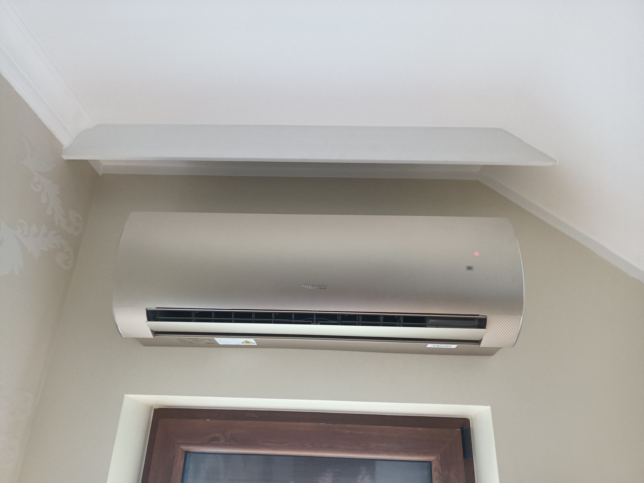 Zamontowany klimatyzator w jasnym wnętrzu, widoczny pod sufitem, z dekoracyjną listwą. Ściana w delikatny wzór, dół kadru okno z brązową ramą.