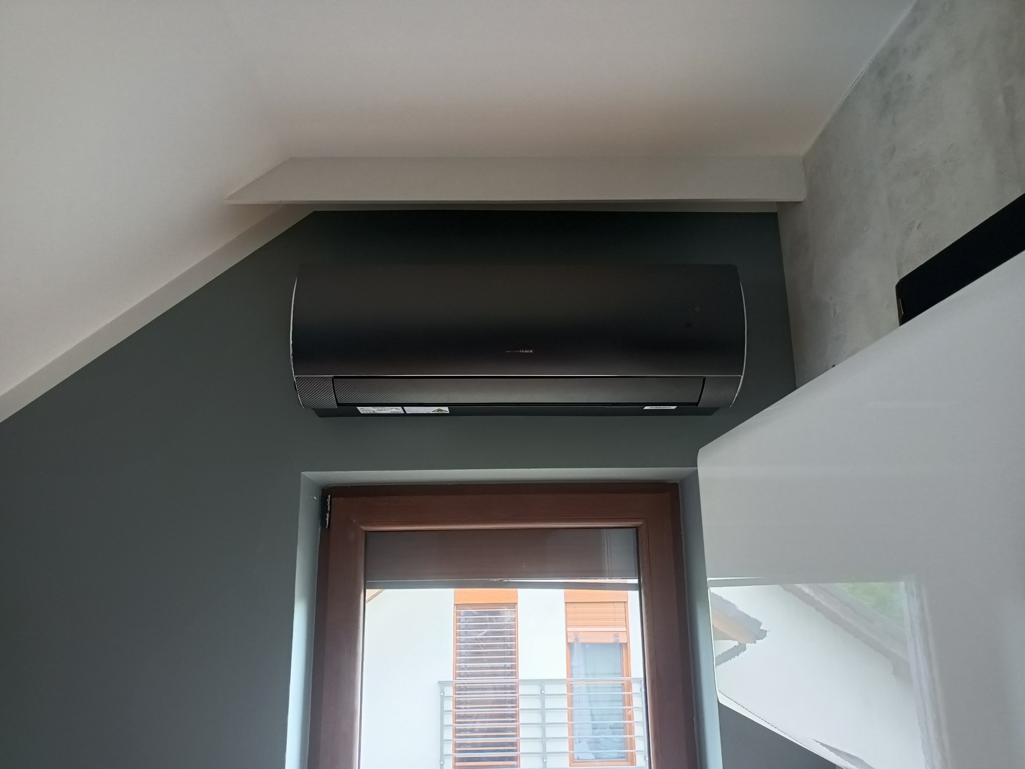 Czarny klimatyzator ścienny zamontowany nad oknem z brązową ramą w szaro-białym wnętrzu domu. Widoczne elementy zabudowy.