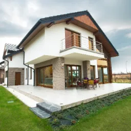 Nowoczesny dom murowany z drewnianym wykończeniem dachu, balkonem z metalową balustradą i przestronnym tarasem z meblami ogrodowymi, otoczony zielonym trawnikiem i schodami z kamienia łupanego.