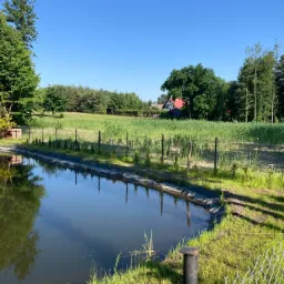 Wykończony zbiornik wodny z folią, otoczony ogrodzeniem z siatki, z widokiem na zarośnięte pole i dom z czerwonym dachem w tle, w słoneczny dzień.