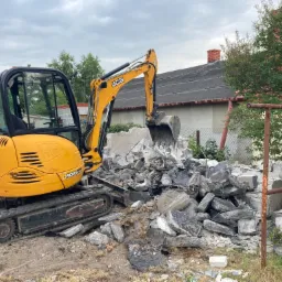 Żółta mini koparka JCB 8025 ZTS z łyżką pełną gruzu i fragmentów betonu na placu budowy z widocznym budynkiem w tle.