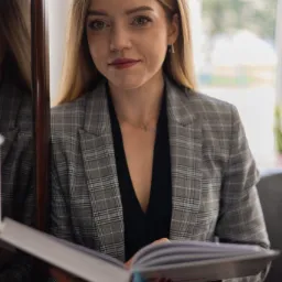 Kobieta w szarym żakiecie w kratę, trzyma otwarta książkę z niebieskimi brzegami kartek, stojąc obok lustra.