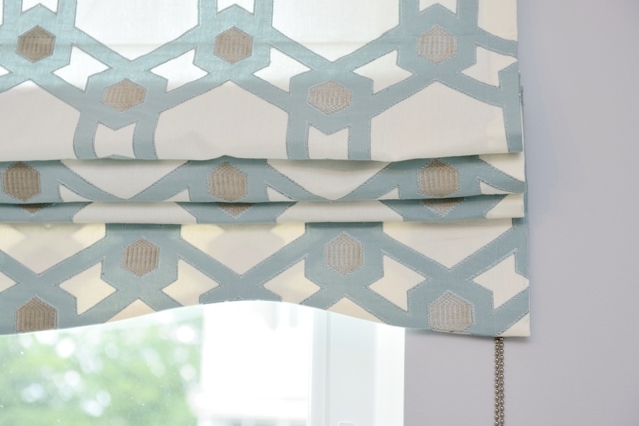 Złożona roleta rzymska w kolorze biało-błękitnym z geometrycznym wzorem, zamontowana w oknie, z widocznym łańcuszkiem sterującym.