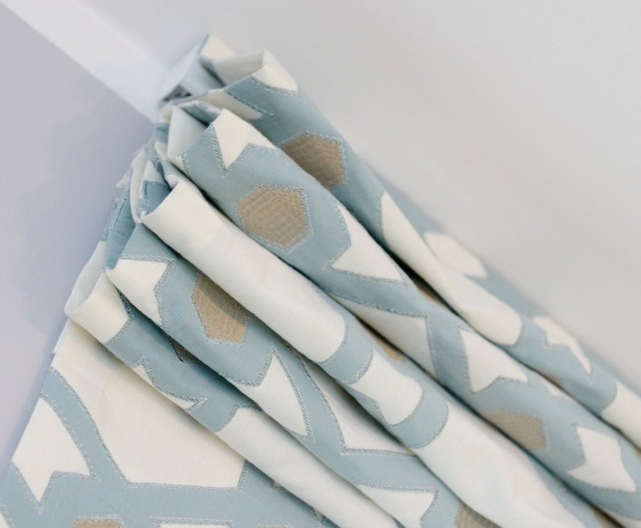 Złożona, pastelowa roleta okienna z geometrycznym wzorem w odcieniach błękitu, beżu i bieli, zamontowana na karniszu.