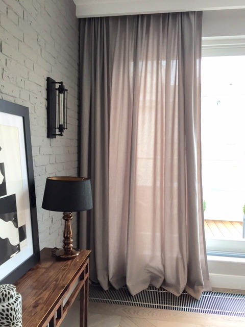 Jasnoszare, delikatne zasłony z subtelnym marszczeniem, wiszące obok okna z widokiem na balkon, uzupełnione przez lampkę nocną na drewnianym stoliku i ceglaną ścianę z kinkietem.