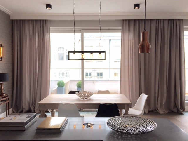 Eleganckie, długie, beżowe zasłony w nowoczesnym salonie z wyjściem na balkon, oświetlone wiszącymi lampami i światłem dziennym. Widoczne elementy dekoracyjne na stole i para książek.