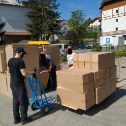 Pracownicy w maskach ładują kartonowe pudła na niebieski wózek transportowy na zewnątrz budynku, w tle budynki mieszkalne i parking.