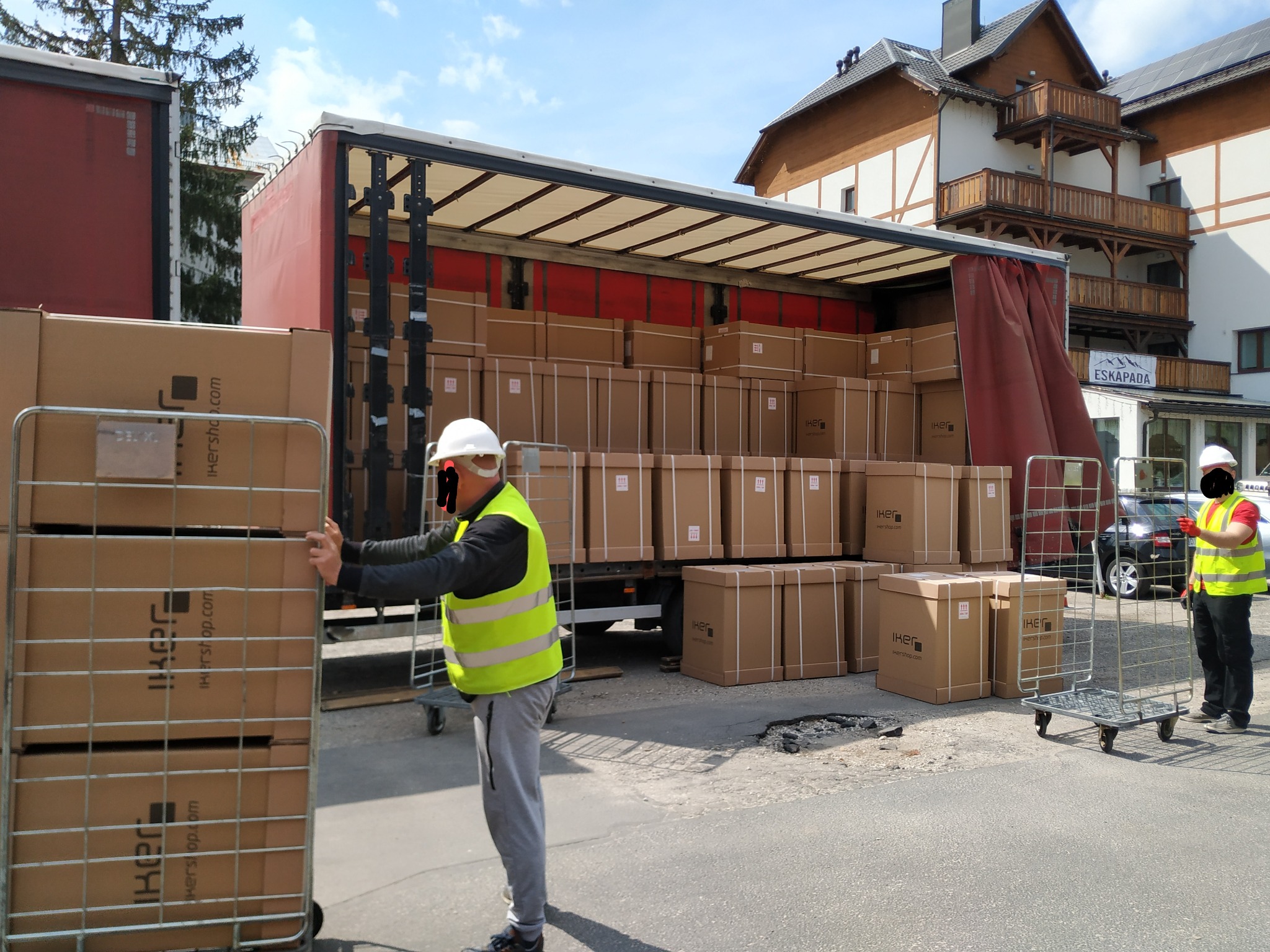 Pracownicy w kamizelkach odblaskowych rozładowują z ciężarówki kartony z logo IKERSHOP na wózkach transportowych, budynek z balkonami w tle.