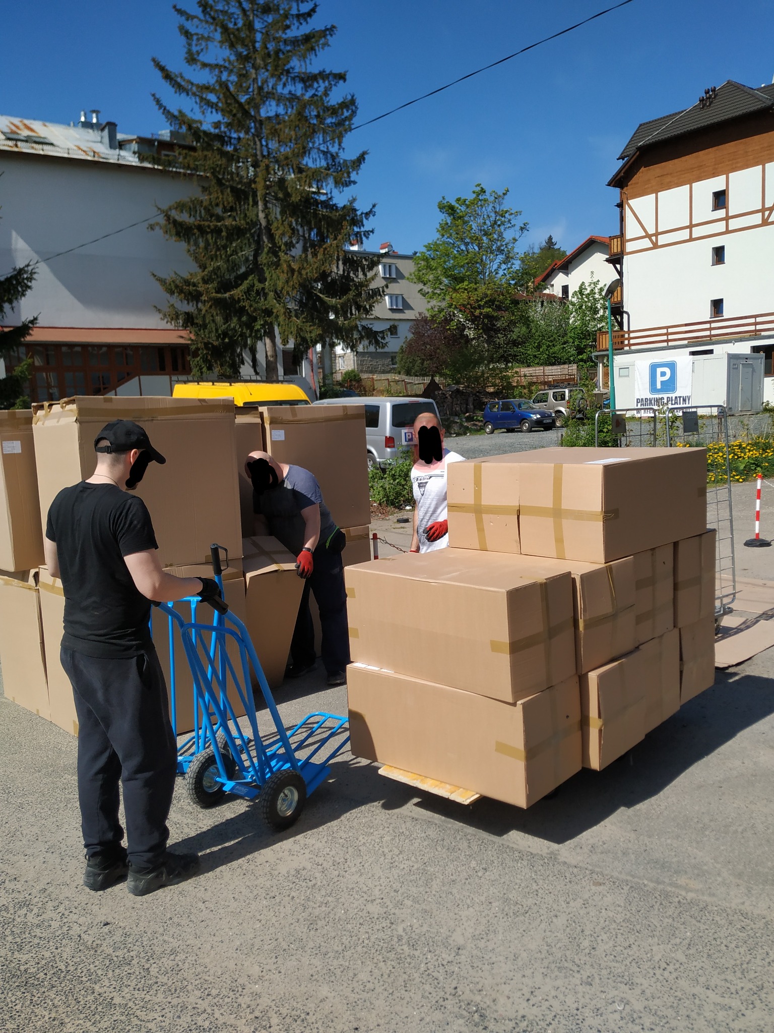 Pracownicy w maskach ładują kartonowe pudła na niebieski wózek transportowy na zewnątrz budynku, w tle budynki mieszkalne i parking.