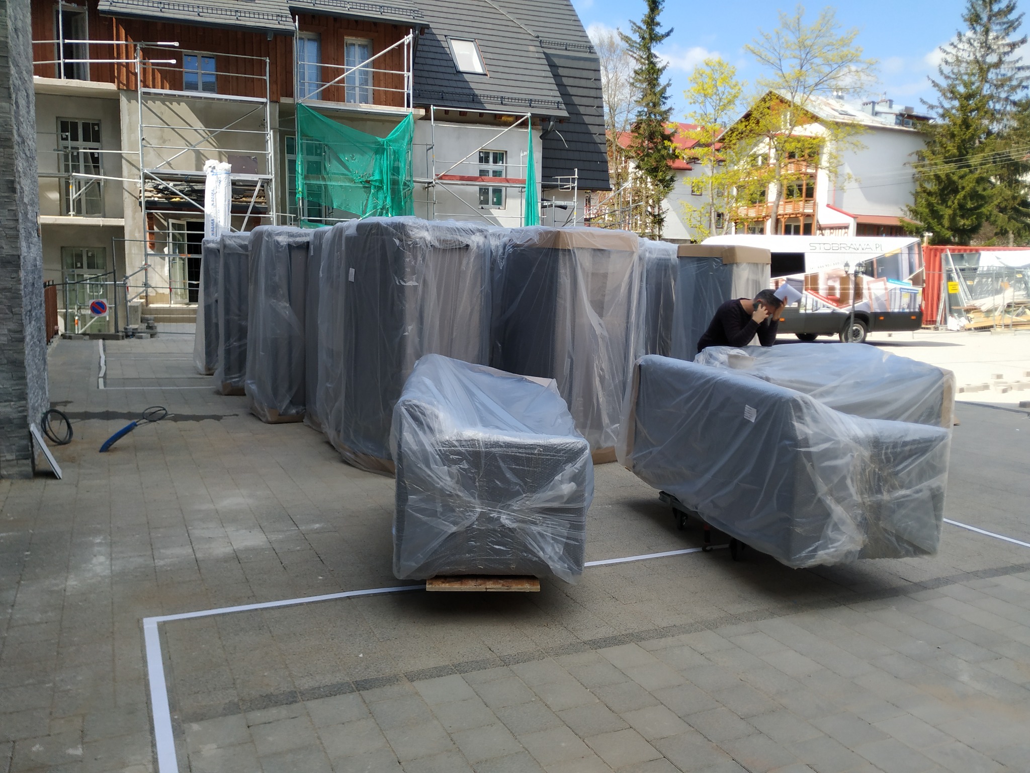 Meble zapakowane w folię ochronną, przygotowane do transportu, na tle budynku w trakcie budowy, z widocznym rusztowaniem i busem z napisem 'Stobrawa.pl'.