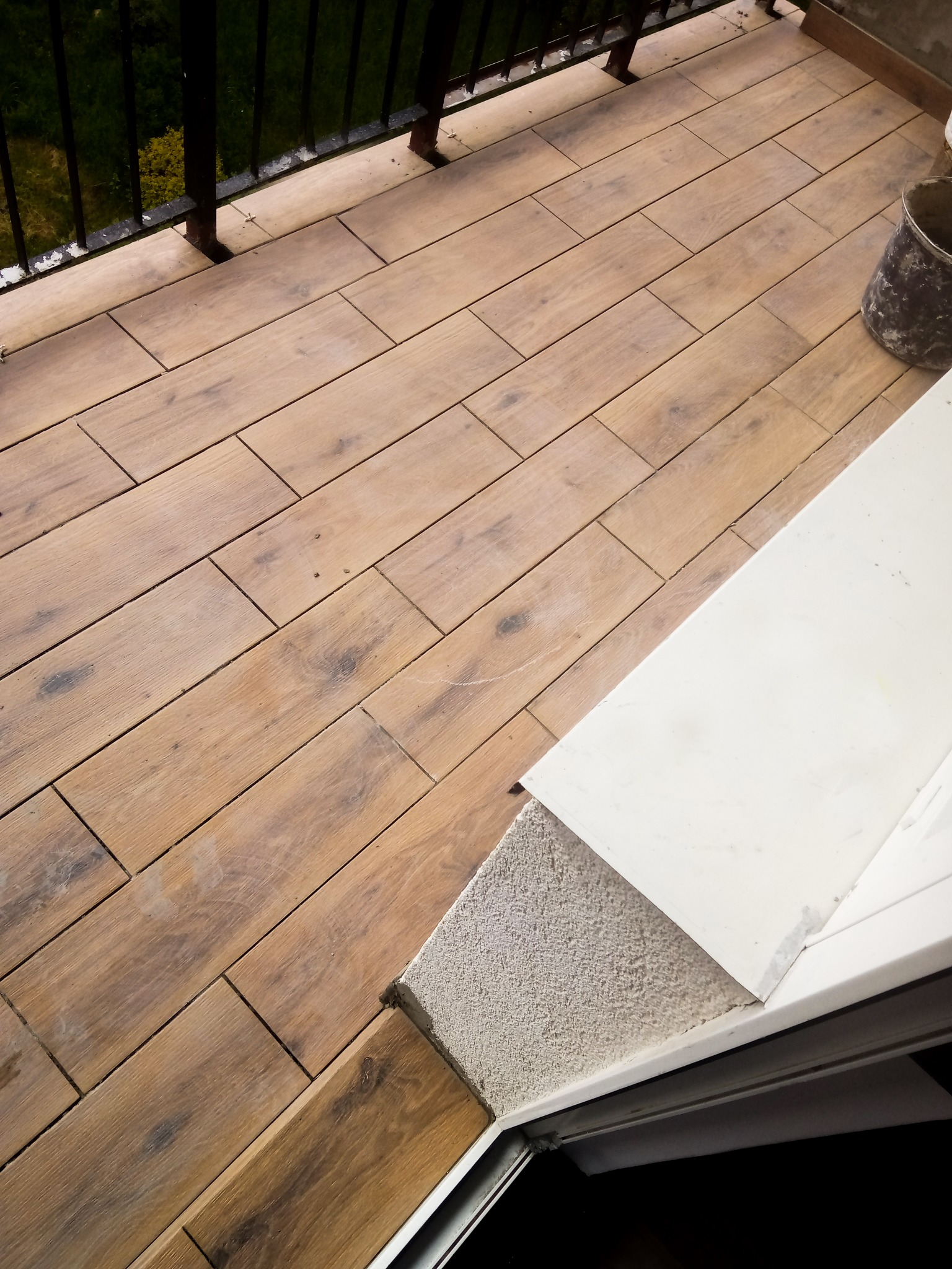 Ułożona pod kątem brązowa terakota imitująca drewno na balkonie, widoczny fragment balustrady, biały próg i wiaderko z zaprawą.