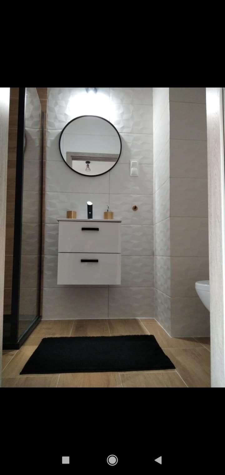 Nowoczesna łazienka z umywalką nablatową, okrągłym lustrem w czarnej ramie, płytkami 3D na ścianie i drewnopodobną podłogą, uzupełniona czarnym dywanikiem.