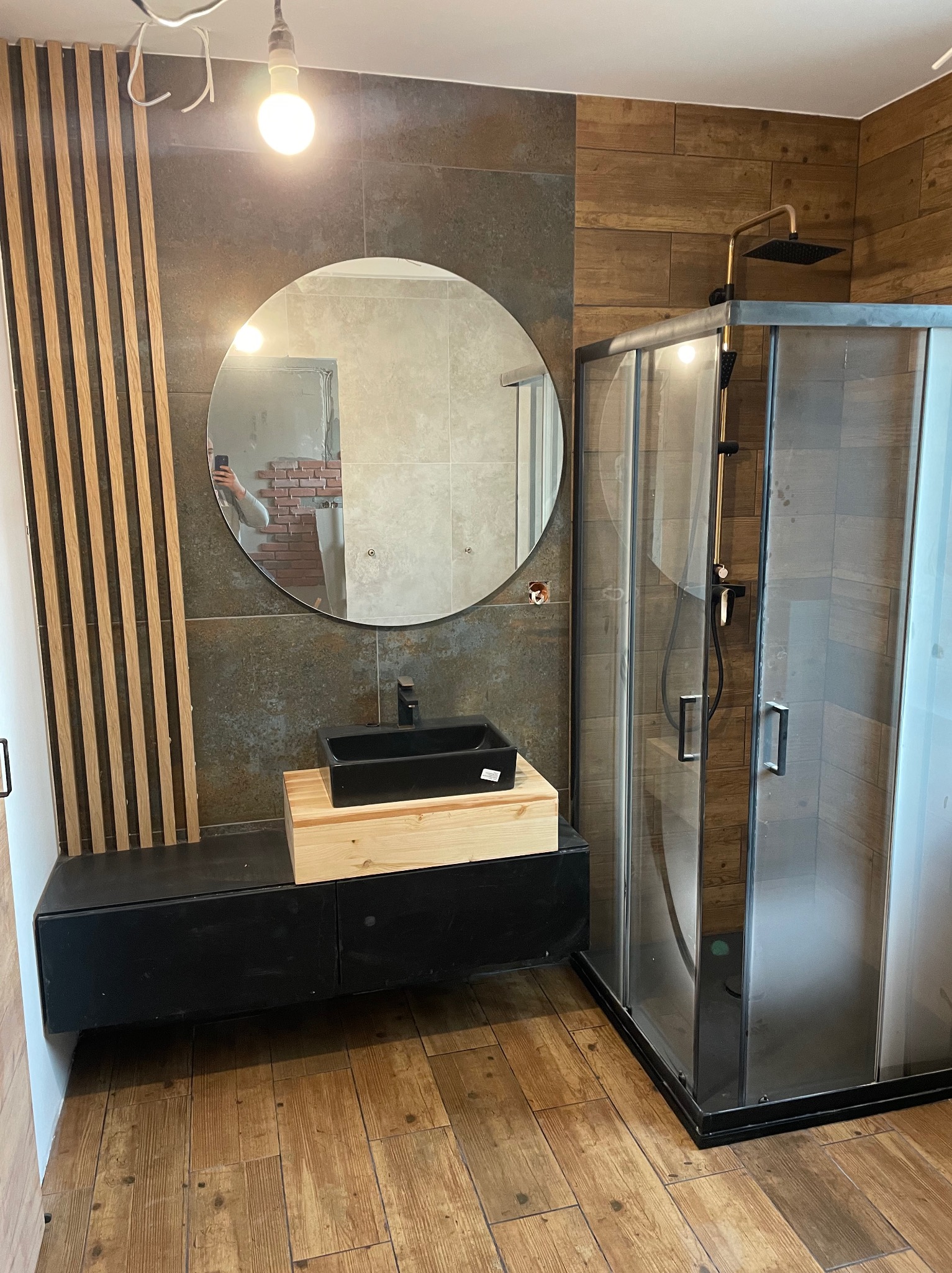 Nowoczesna łazienka z drewnianymi akcentami: kabina prysznicowa z czarnymi ramami, okrągłe lustro z widocznym odbiciem części ściany z cegły, czarna umywalka na drewnianej podstawie, drewniane...