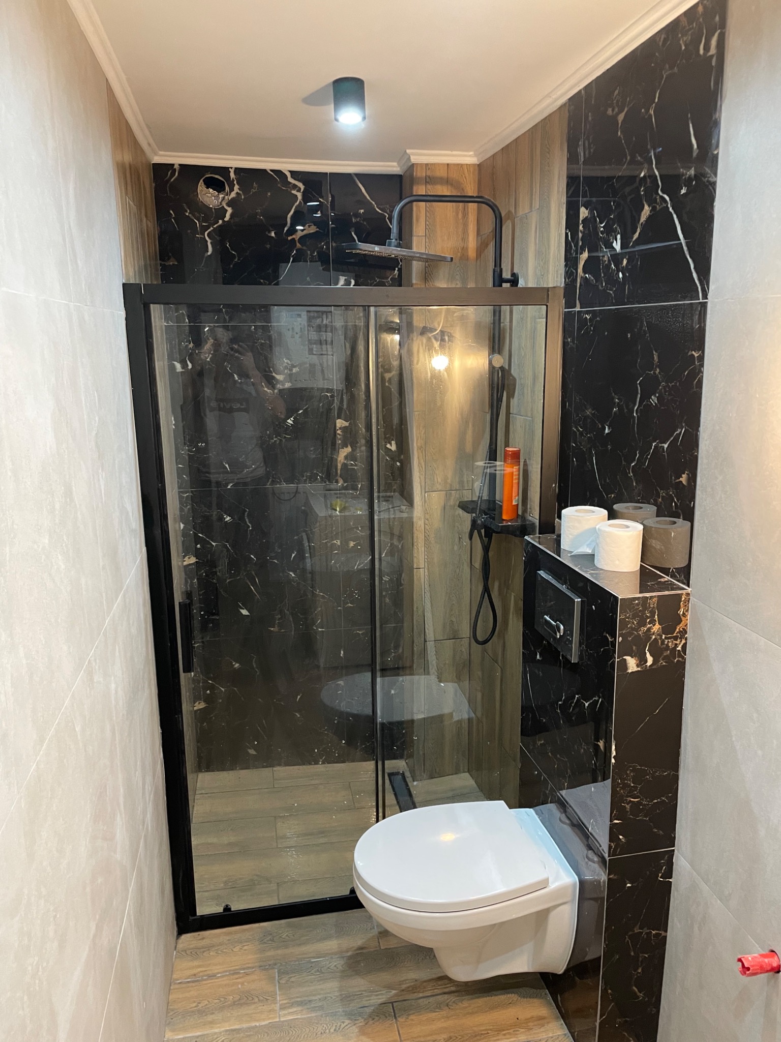 Nowoczesna łazienka z kabiną prysznicową typu walk-in, płytki imitujące czarny marmur z białymi żyłami, podwieszana toaleta i szare płytki na ścianach.