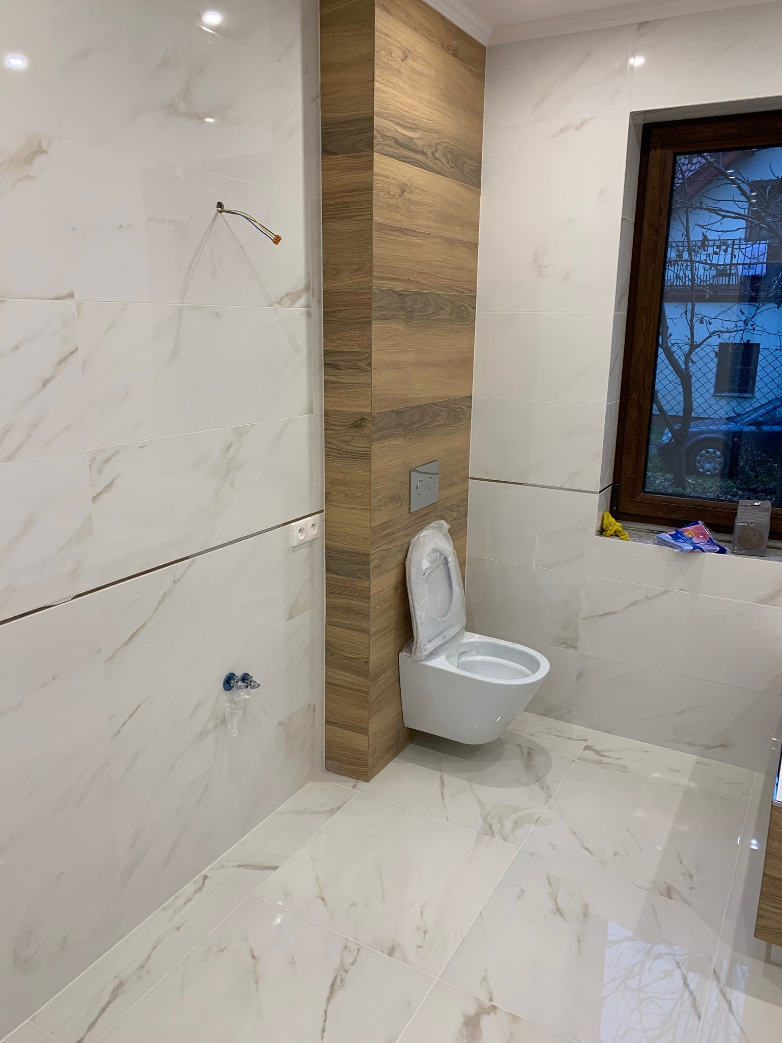 Nowoczesna łazienka w trakcie wykańczania: białe płytki imitujące marmur na ścianach i podłodze, pionowy panel z drewnopodobnych płytek, wisząca toaleta zabezpieczona folią, widoczne przyłącza...