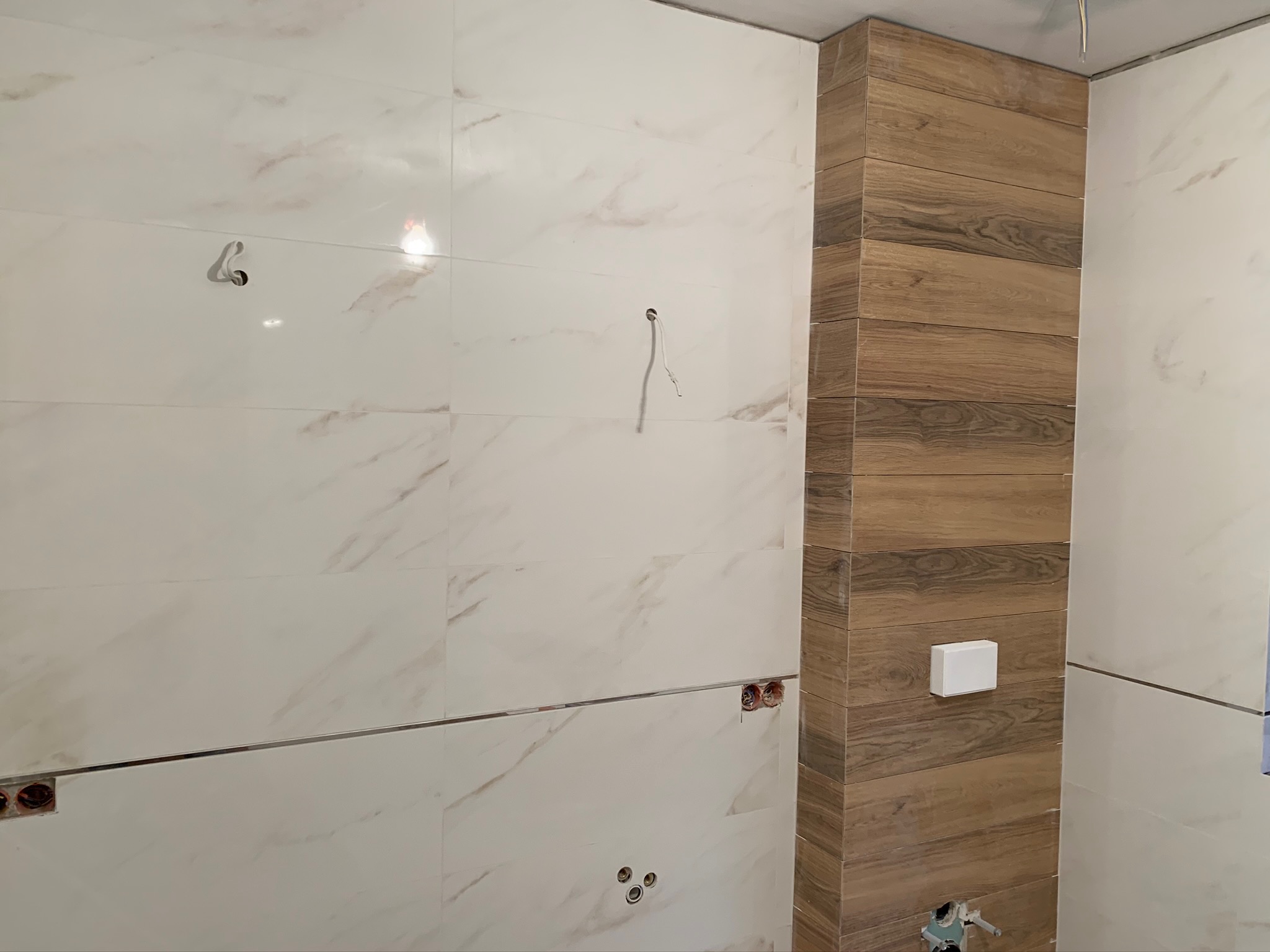 Ściana łazienki w trakcie wykańczania, z białymi płytkami imitującymi marmur i kolumną wykończoną płytkami drewnopodobnymi, widoczne puszki elektryczne i wyprowadzenia hydrauliczne.