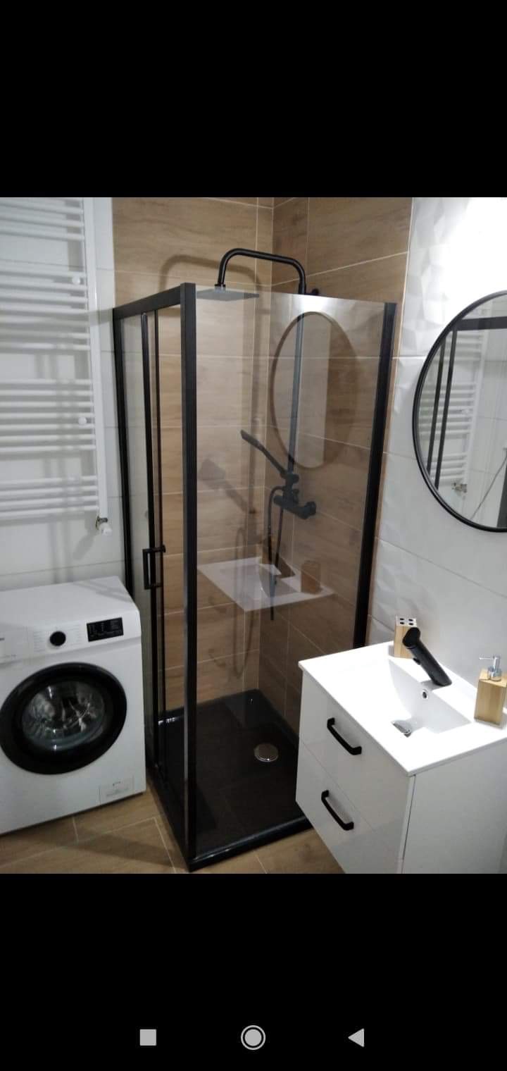 Nowoczesna łazienka z kabiną prysznicową typu walk-in z czarnymi profilami, białą szafką z umywalką i pralką, wykończona płytkami imitującymi drewno.