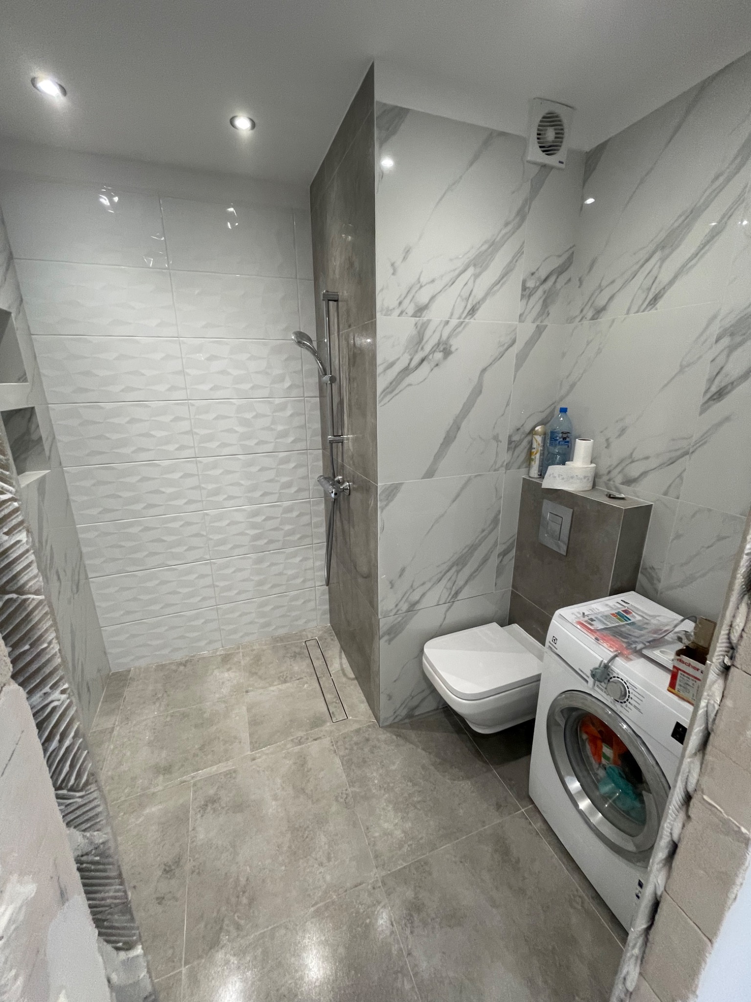 Wykończona łazienka z szarymi płytkami imitującymi marmur na ścianach, białe płytki 3D pod prysznicem, szara podłoga, biała toaleta i pralka Electrolux, widoczny fragment ściany w trakcie prac...
