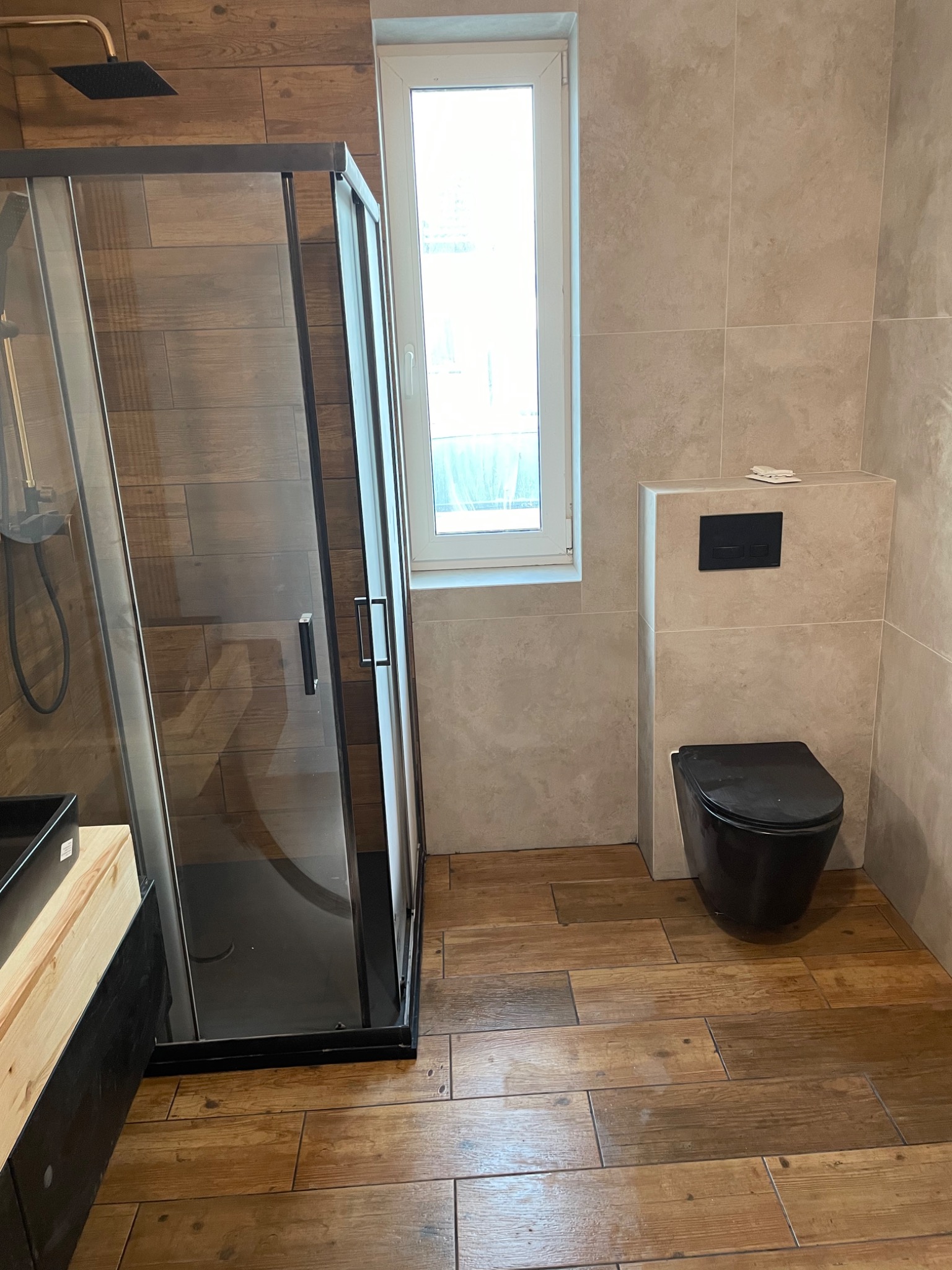 Nowoczesna łazienka z kabiną prysznicową typu walk-in, czarną armaturą, podwieszaną toaletą w kolorze czarnym i podłogą wyłożoną płytkami imitującymi drewno.