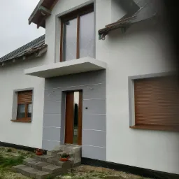 Dwukondygnacyjny dom z jasną elewacją, brązowymi roletami i drewnianymi drzwiami wejściowymi, widoczne schody z betonowych bloków i doniczki z kwiatami.