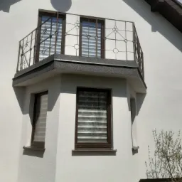 Biała elewacja domu z narożnym balkonem z metalową balustradą o dekoracyjnym wzorze, widoczne okna z brązowymi ramami i roletami dzień-noc.