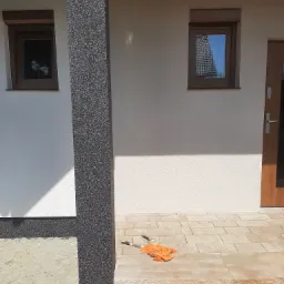 Wykończona elewacja domu z kolumną w ciemnym, ziarnistym tynku, jasną elewacją, oknami z brązowymi ramami i schodami wejściowymi z kamiennej kostki, na których leżą pędzle malarskie.