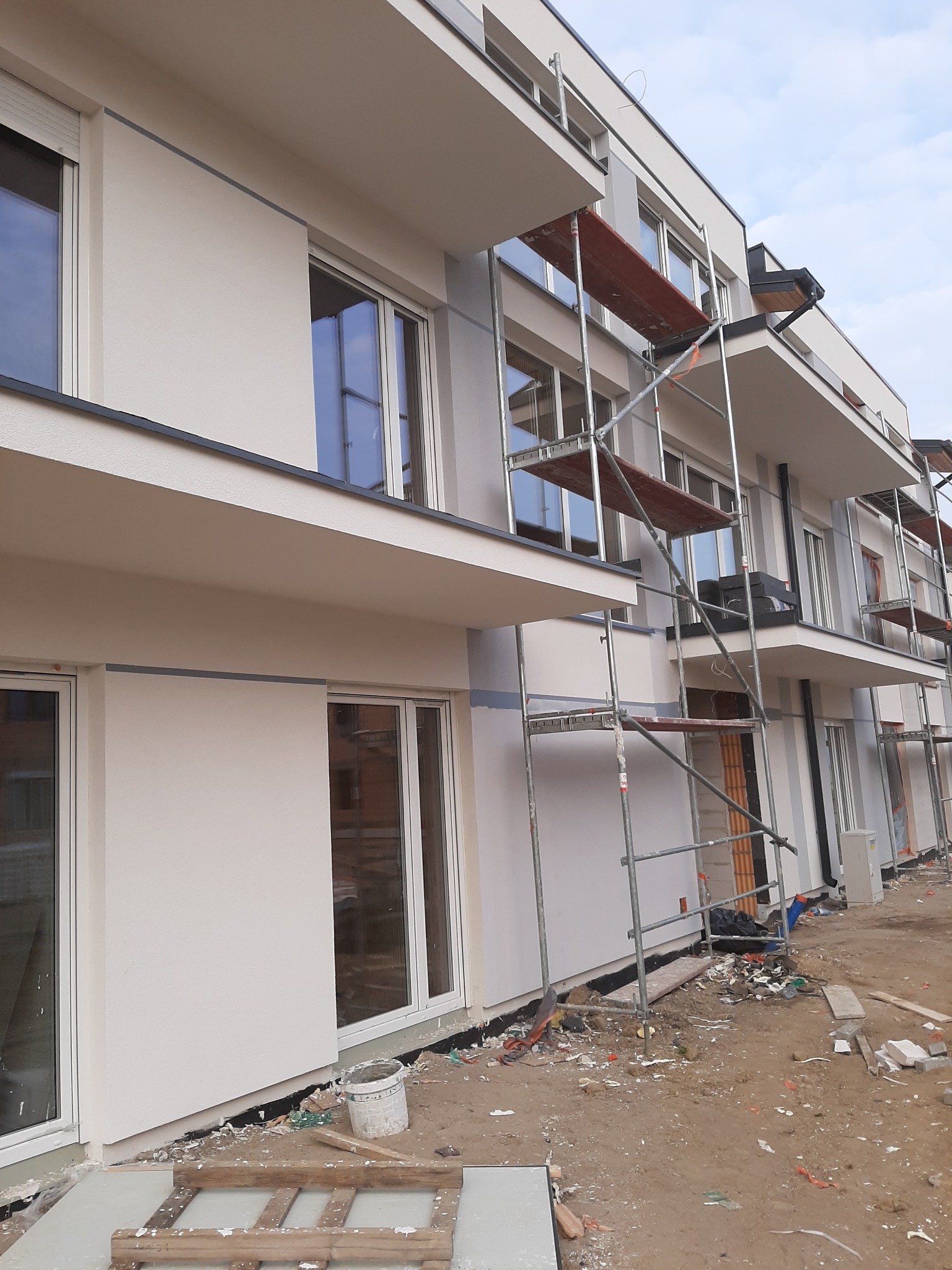 Nowoczesny budynek wielorodzinny z ocieploną i otynkowaną elewacją w kolorze jasnego beżu, z balkonami i rusztowaniami podczas prac wykończeniowych.