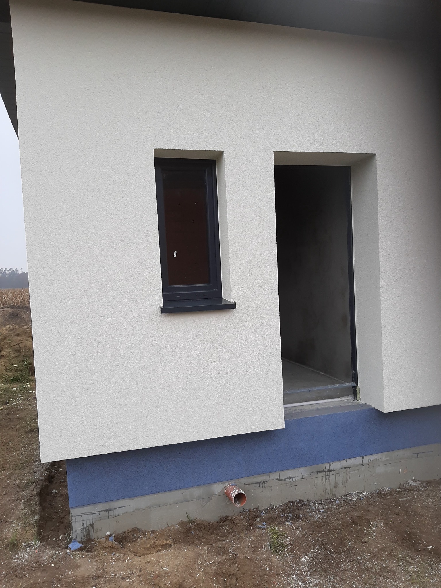 Fragment elewacji budynku z oknem i otworem drzwiowym w stanie surowym, widoczna struktura tynku i cokół w kolorze niebieskim, teren wokół budynku niezagospodarowany.