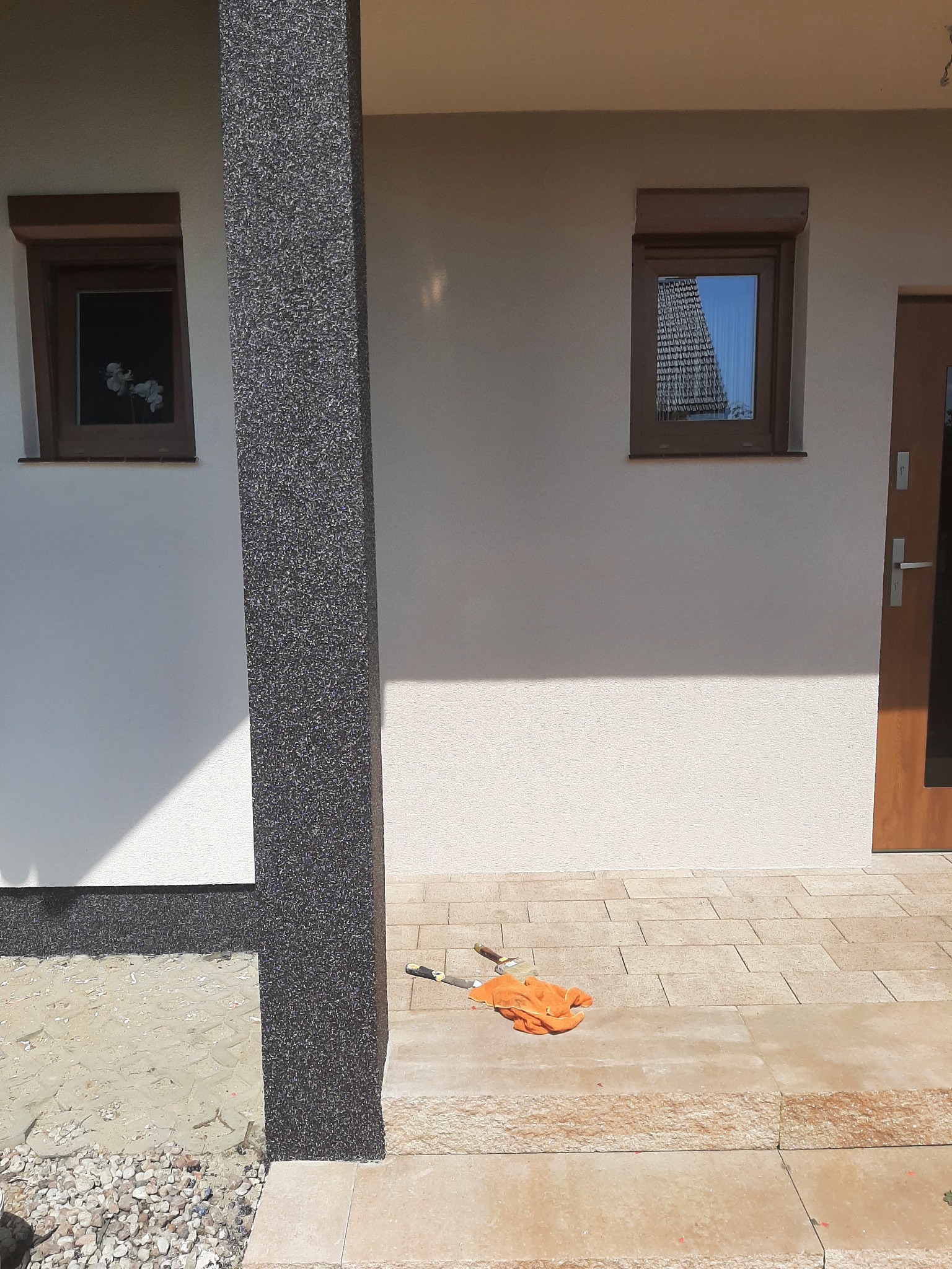 Wykończona elewacja domu z kolumną w ciemnym, ziarnistym tynku, jasną elewacją, oknami z brązowymi ramami i schodami wejściowymi z kamiennej kostki, na których leżą pędzle malarskie.