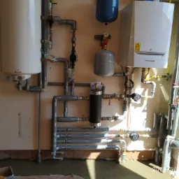 Kompleksowa instalacja grzewcza z kotłem De Dietrich, zasobnikiem Galmet i licznymi rurami, widoczny etap montażu z resztkami zaprawy na podłodze i łopatą w tle.