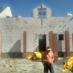 R.P.S-Studio - Wznoszenie ścian domu z bloczków betonowych i cegieł, widoczny murarz w pomarańczowej koszulce i czapce, teren budowy z piaskiem i gruzem.