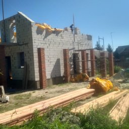 R.P.S-Studio - Budowa domu w Łodzi: surowy stan z białych bloczków, częściowo obłożony czerwoną cegłą, drewniane stemple, widoczne rusztowania i składowane deski na tle błękitnego nieba.