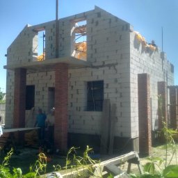 R.P.S-Studio - Surowy stan budynku w trakcie wznoszenia, ściany z betonu komórkowego i ceglane filary, widoczni dwaj pracownicy oraz narzędzia budowlane na tle zieleni.