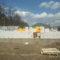 R.P.S-Studio - Wznoszenie muru z białych bloczków, częściowo zabezpieczonych żółtą folią, na fundamencie; w tle widoczny pracownik w zielonej koszuli, drzewa i zabudowania mieszkalne.