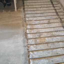 R.P.S-Studio - Montaż rur ogrzewania podłogowego na siatce w surowym pomieszczeniu, widoczne niedoskonałości podłoża i data na zdjęciu.